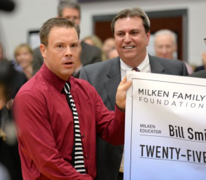 Milken Educator Award recipient Bill Smithyman is too stunned to smile as he stands in front of the packed gym at Blue Valley Northwest High School, where students, faculty and guests stand cheering. Kansas Commissioner of Education Dr. Randy Watson looks on with a smile. photo Milken Educator Awards